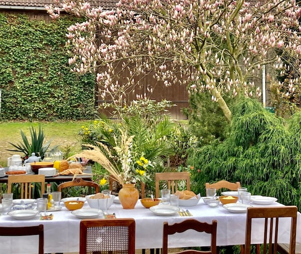 An outdoor dining area with a table set for a meal, surrounded by greenery and flowering trees. - "Hühnerstudio - Schlamau19" Studio with Shared Terrace and Shared Garden  (Wiesenburg/Mark)