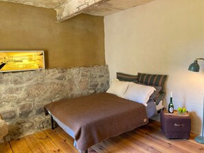 A bedroom with a stone wall, a bed with a brown blanket, a nightstand with a bottle and apples, and a lamp. - "Hühnerstudio - Schlamau19" Studio with Shared Terrace and Shared Garden  (Wiesenburg/Mark)