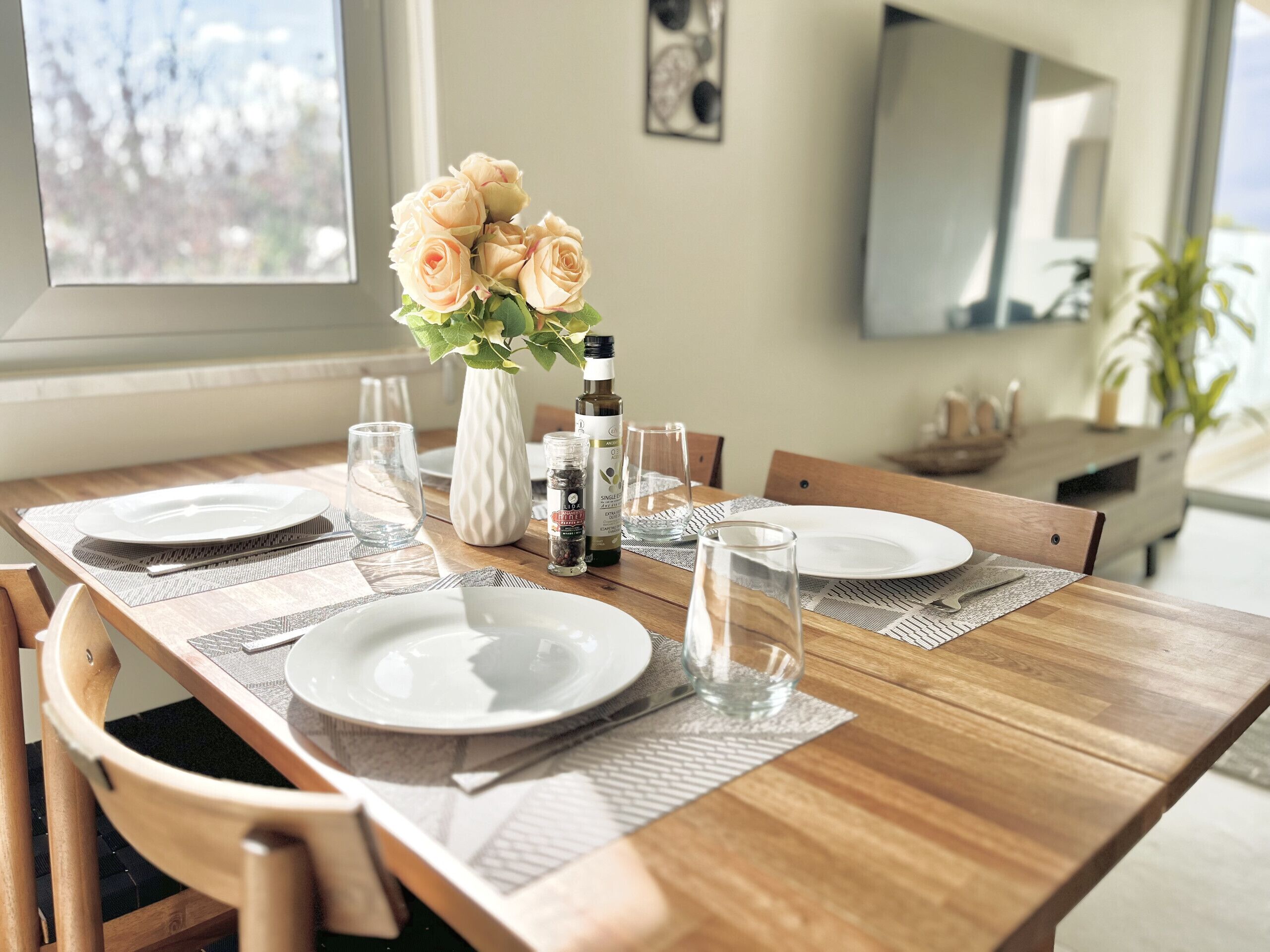 Een eettafel gedekt met borden, glazen, een vaas met bloemen en een fles olijfolie.