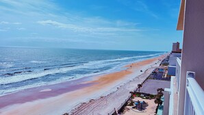 Vista a la playa desde un balcón, cielo despejado, arena y algunos edificios al fondo.