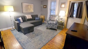 A living room with a grey sofa, armchair, and ottoman, a patterned rug, and a wooden floor.