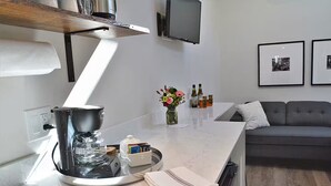 Une cuisine moderne équipée d’une machine à café, d’un petit évier et d’un plan de travail agrémenté d’un vase de fleurs. Le salon comprend un canapé et un téléviseur à écran plat.