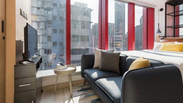 Una habitación de hotel moderna con un ventanal grande, un sofá, una mesita y un televisor.