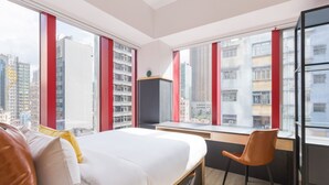 Una habitación de hotel moderna con cama, escritorio y silla, ventanas amplias con vistas a la ciudad y un paisaje urbano visible a través de las ventanas.
