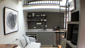 A compact kitchen with a built-in oven, a microwave, and a coffee maker. There is a grey sofa with a throw blanket and a framed artwork on the wall. A TV is mounted on the wall above the kitchen. - The Wiedler - Charming Tiny Home - AC, WiFi in the heart of the action! (Portland)