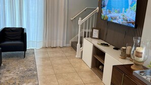 Un salon moderne doté d’un grand téléviseur à écran plat fixé sur un panneau en bois, d’un canapé en cuir noir, d’un escalier avec des rampes blanches et d’une vue sur la mer à travers des rideaux légers.