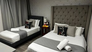 Une chambre d’hôtel avec deux lits, chacun doté d’une tête de lit grise, de draps blancs et d’oreillers à l’effigie de Mickey Mouse. On y trouve une petite table avec une lampe et une figurine de Mickey Mouse.