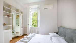 A bedroom with a bed, a grey upholstered headboard, a window with a view of greenery, and a white wardrobe with open doors. - Family stay in Trastevere (Rome)