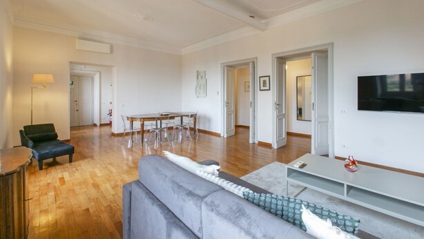 A spacious living area with a grey sofa, a wooden dining table with chairs, a TV mounted on the wall, and a lamp on a side table. - Family stay in Trastevere (Rome)