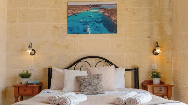A neatly made bed with pillows and rolled towels, a painting of a coastal scene on the wall, and two wall-mounted light fixtures. - Ta Dora Sleeps 6 with Private Pool (Xaghra)