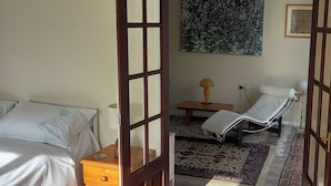 Een kamer met een bed, nachtkastje, stoel, lamp, vloerkleed en een schilderij aan de muur.