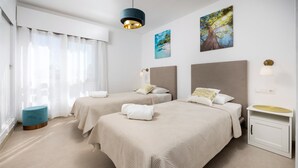 A hotel room with two beds, a small round ottoman, a bedside table with a lamp, and a wall-mounted shelf. - Estepona Beachfront Promenade by Libehomes (Estepona)