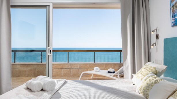 A hotel room with a bed, a balcony with a view of the sea, and a chair with a hat and towel. - Estepona Beachfront Promenade by Libehomes (Estepona)