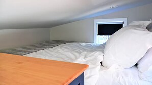 A small, well-lit room with a single bed, a wooden table, and a window with a black blind.