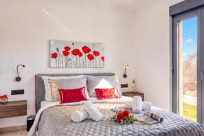 A modern bedroom with a large bed, two nightstands, a wall-mounted lamp, and a window with a view.