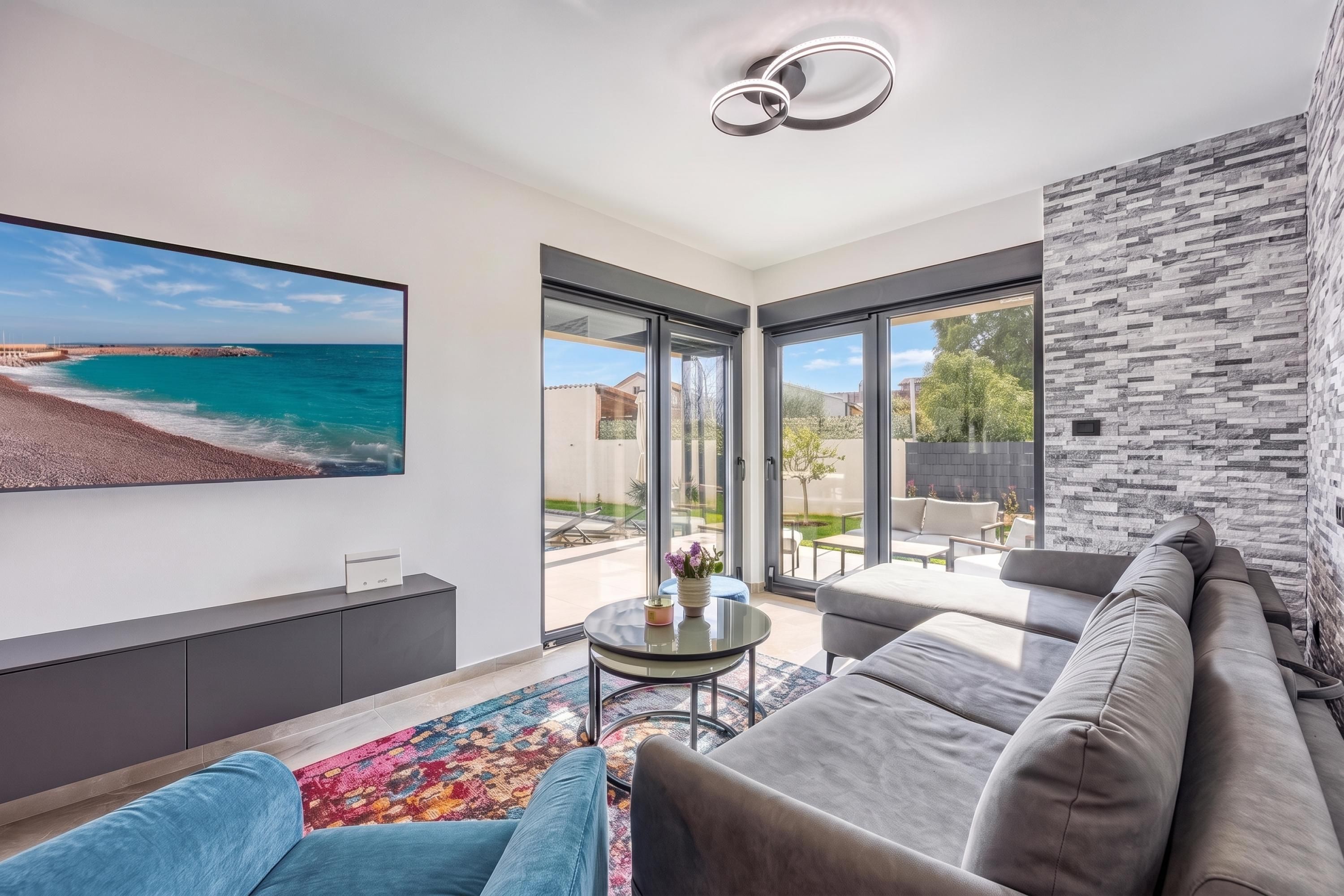 A modern living room with a large TV, comfortable seating, and a view of the sea.
