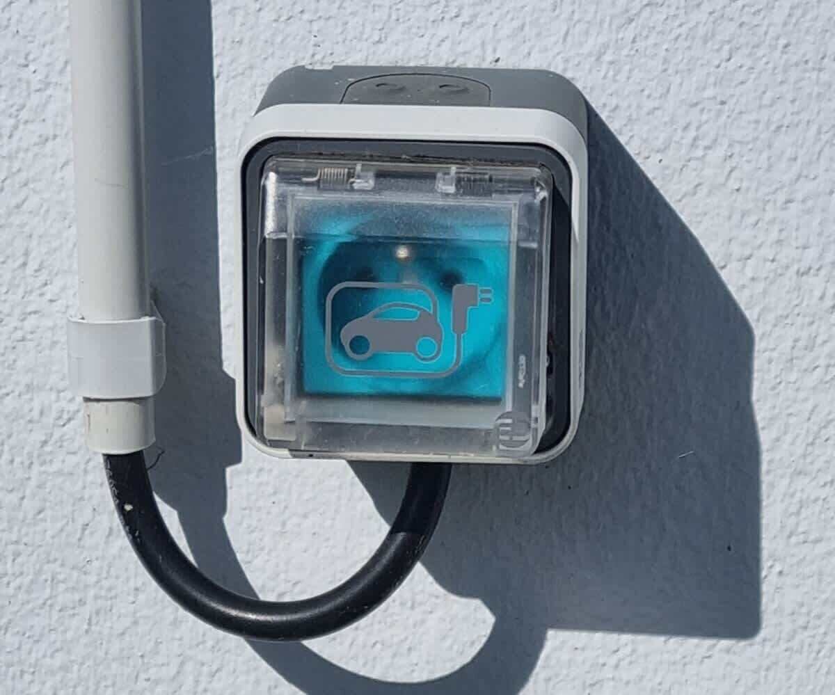 Uma estação de carregamento portátil para veículos elétricos, com símbolo de carregamento visível, um cabo e uma caixa fixada na parede.