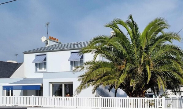 Un bâtiment à deux étages avec une façade blanche, une antenne parabolique et une clôture en bois peinte en blanc.