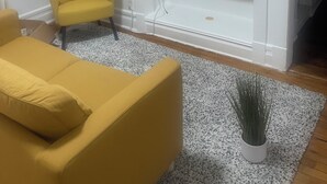 Una sala de estar con un sofá amarillo, un televisor sobre un estante blanco y una planta pequeña sobre una alfombra.