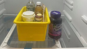 Un refrigerador vacío con un recipiente amarillo que contiene una botella de yogur, un frasco de jalea y una botella de agua.