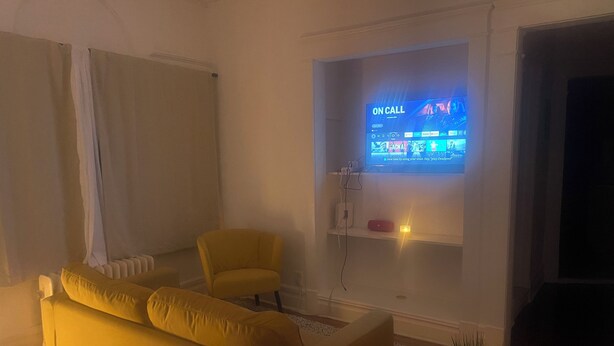 Una sala de estar con un sofá amarillo, un sillón amarillo pequeño y un televisor en la pared mostrando una pantalla con la leyenda 'ON CALL'.