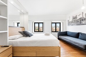 A modern hotel room with a bed, a sofa, a wooden nightstand, and two windows offering city views. - GuestReady - Cathedral Design Apartments - Batalha (Porto)