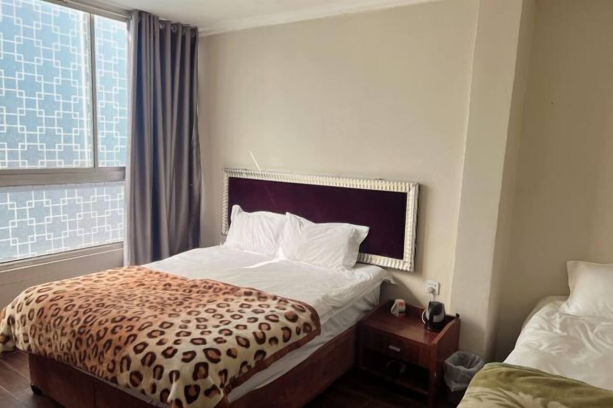 Ein Hotelzimmer mit einem Bett, einem Nachttisch, einem Fenster mit Vorhängen und Blick auf ein Gebäude.