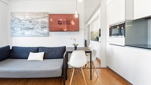 A modern living room with a gray sofa, a dining table with chairs, a potted plant, and a kitchen area with a microwave and oven.