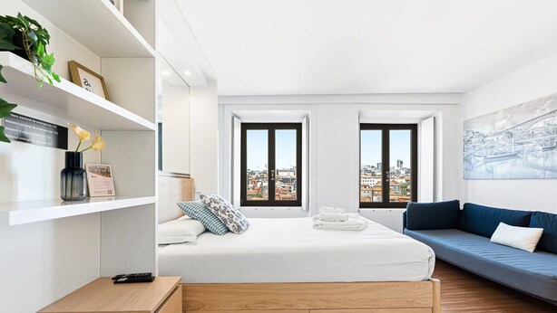 A modern hotel room with a bed, a sofa, a wooden nightstand, a shelf with a vase, and a city view through large windows.