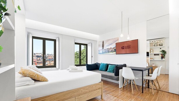 A modern hotel room with a bed, a sofa, a dining area, and a view of the cityscape.