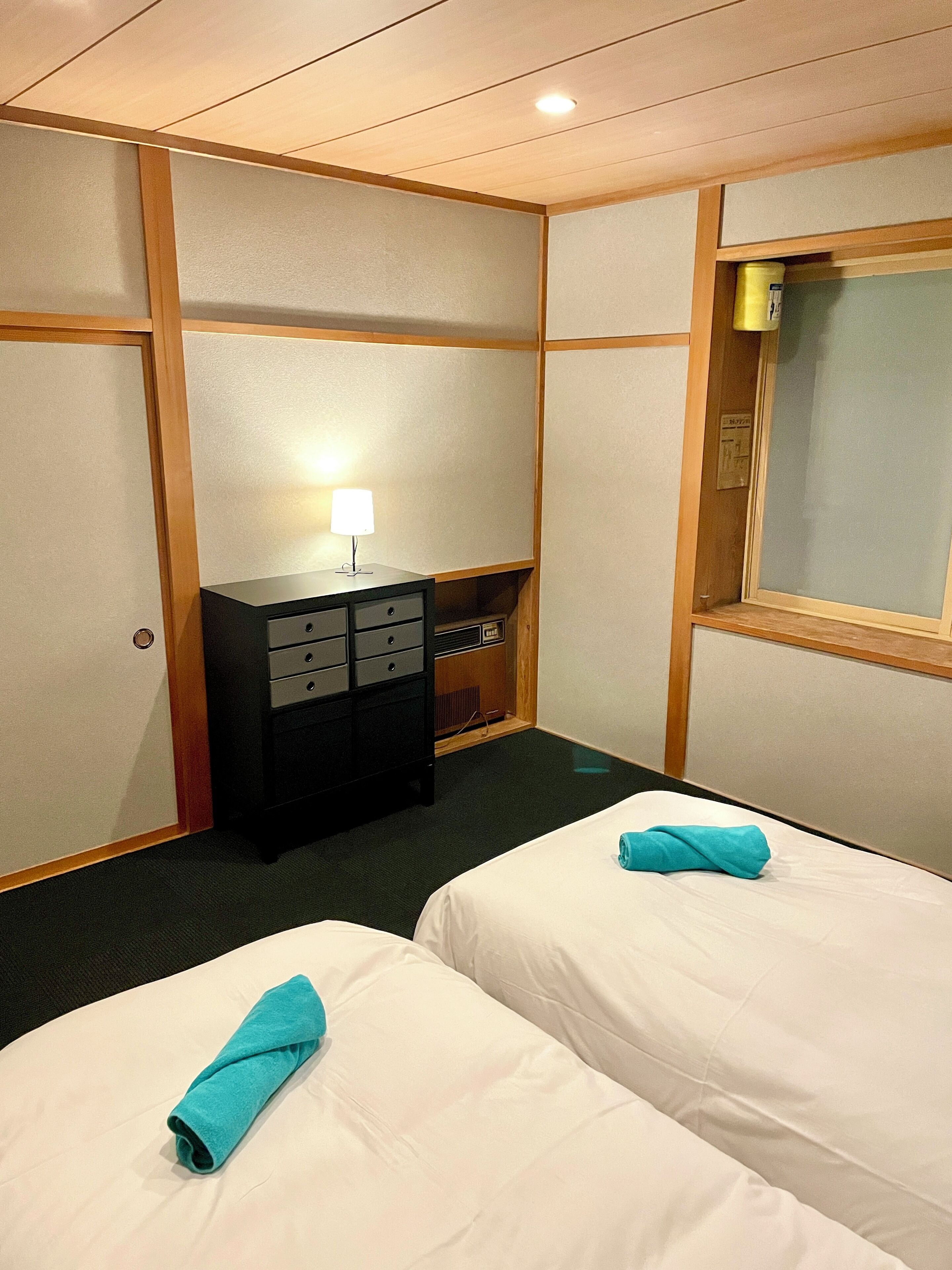 A hotel room with two beds, a desk, a lamp, and a window.