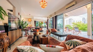 A modern living room with a sofa, coffee table, and TV. There is a large window with a view of buildings and greenery.