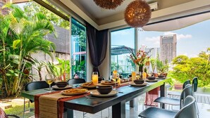 A dining area with a long table set for a meal, surrounded by chairs, with a view of a cityscape through large windows. -  38Pax Flame of the Forest Retreat Villa KL by Verano (Kuala Lumpur)