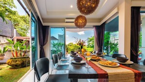A dining area with a long table set for a meal, surrounded by chairs, with a view of greenery and buildings outside.