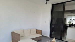 Una sala de estar moderna con un sofá de mimbre y un reposapiés a juego, una puerta de vidrio que da a una zona de cocina y un pequeño objeto decorativo sobre el reposapiés.