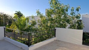 Una terraza en la azotea con barandilla de metal negro, rodeada de vegetación y edificios al fondo.
