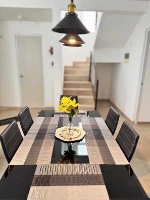 Un comedor con una mesa rectangular, sillas negras, un florero con flores amarillas y una lámpara colgante.
