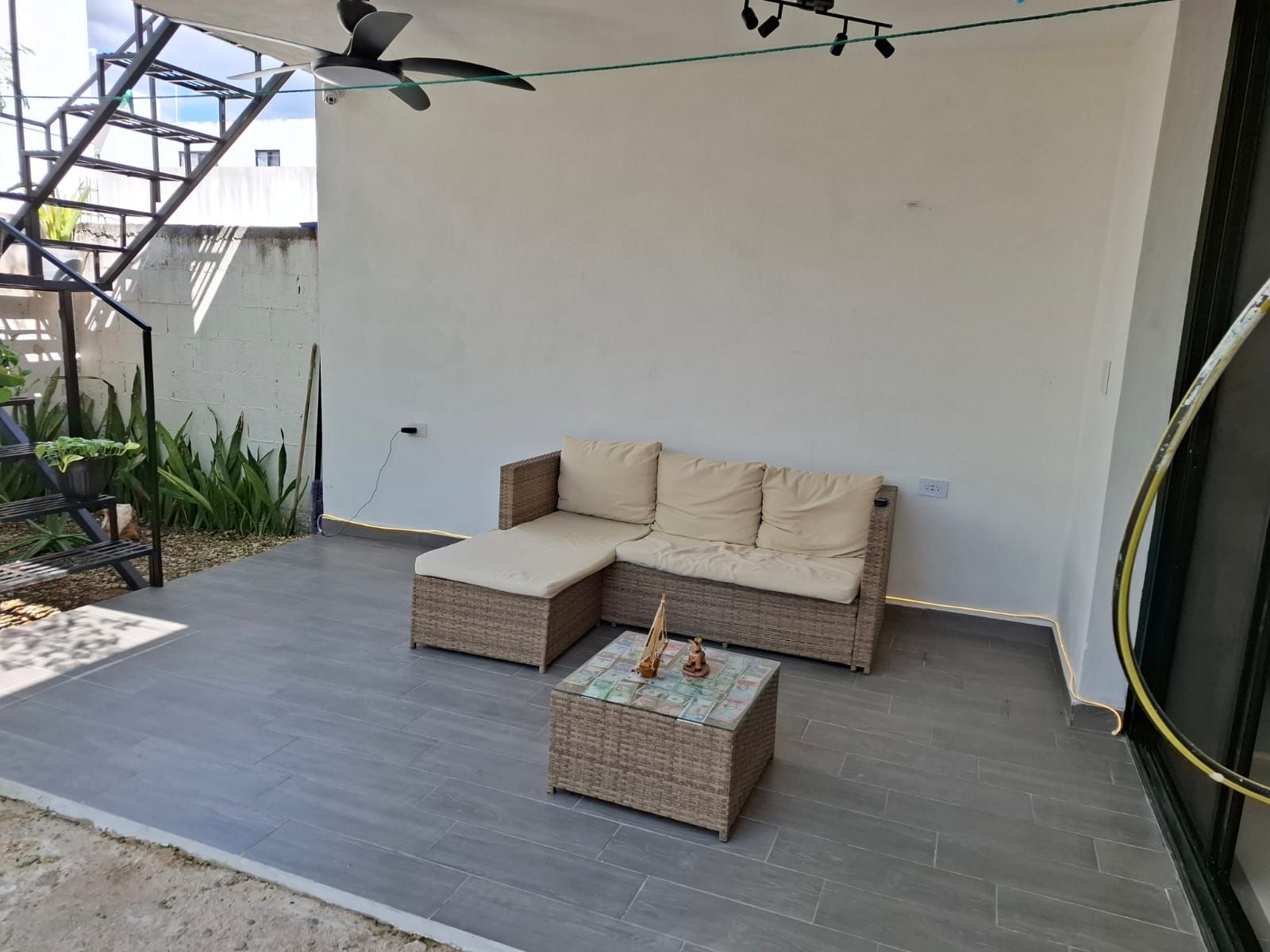 Uma área de estar coberta ao ar livre, com sofá, mesa pequena, ventilador de teto e escada.
