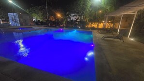 Una piscina con iluminación azul por la noche, rodeada de árboles y una zona cubierta con sillones de descanso.