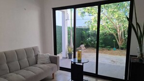Un salón con un sofá gris, una pequeña mesa negra con una caja, y grandes ventanales que dan a un jardín.