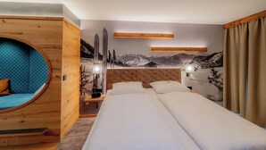 Une chambre d’hôtel dotée d’un grand lit, d’un espace salon circulaire, d’une tête de lit en bois et d’une fresque représentant des montagnes.