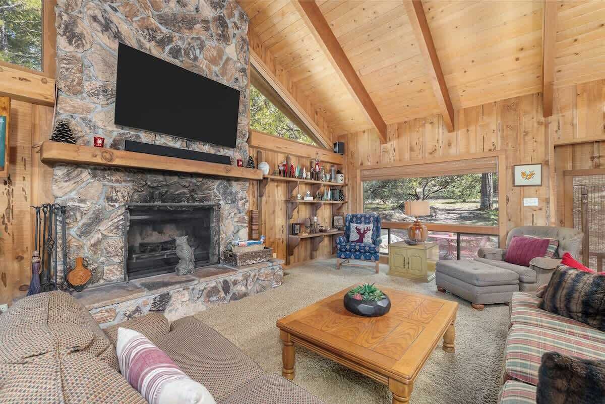 Een knus blokhutinterieur met een stenen open haard, houten planken, een comfortabele zithoek en een groot raam met uitzicht op de natuur.