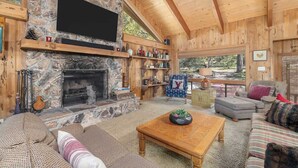 Interior acogedor de una cabaña de troncos con chimenea de piedra, estanterías de madera, una zona de estar confortable y un ventanal con vistas al exterior.