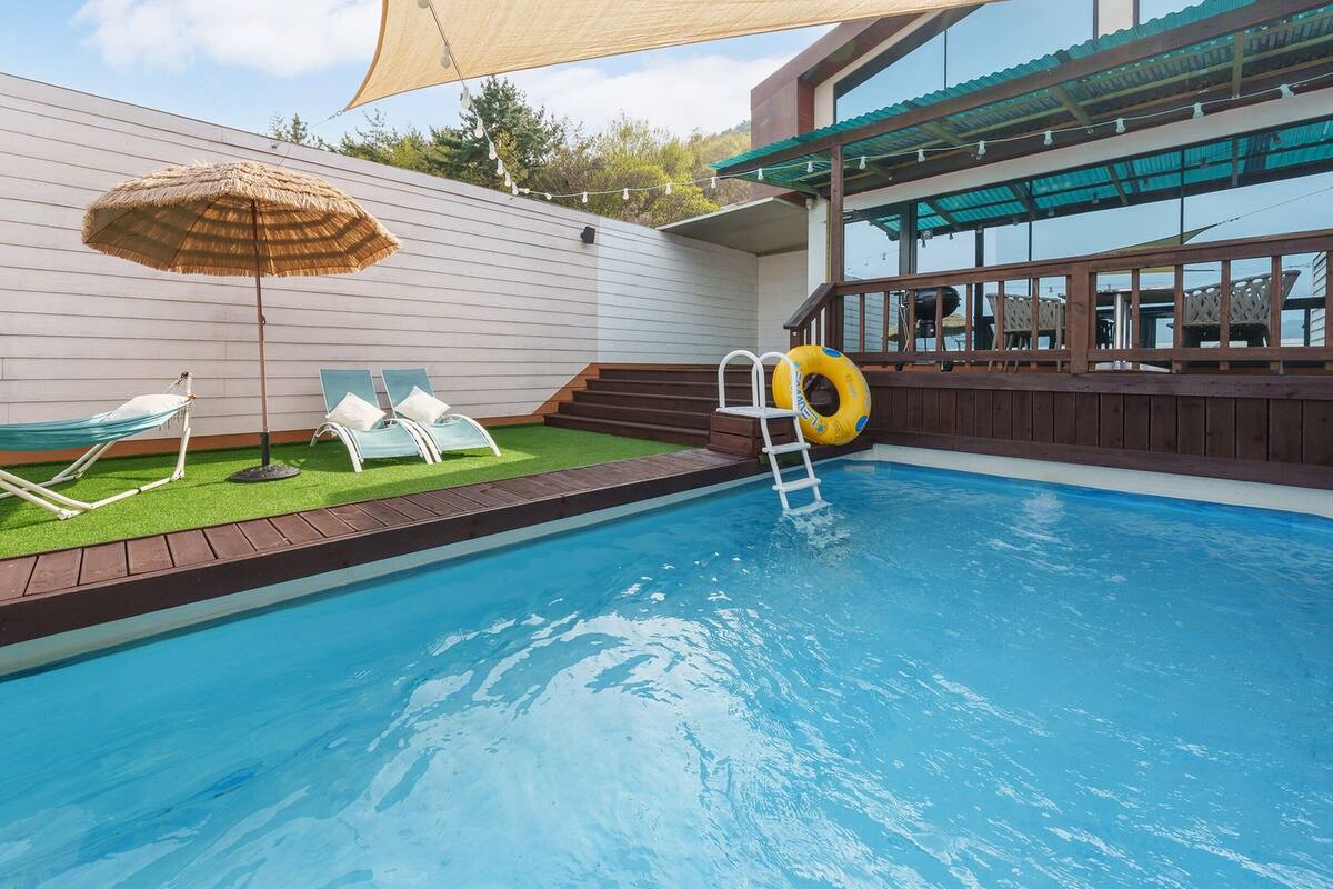 Ein Schwimmbad mit Holzterrasse, Liegestuhl, Hängematte und Sonnenschirm.