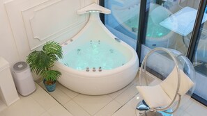 Une salle de bain moderne avec une grande baignoire à remous aux formes arrondies, une plante en pot, une chaise blanche et une douche séparée par des parois en verre.