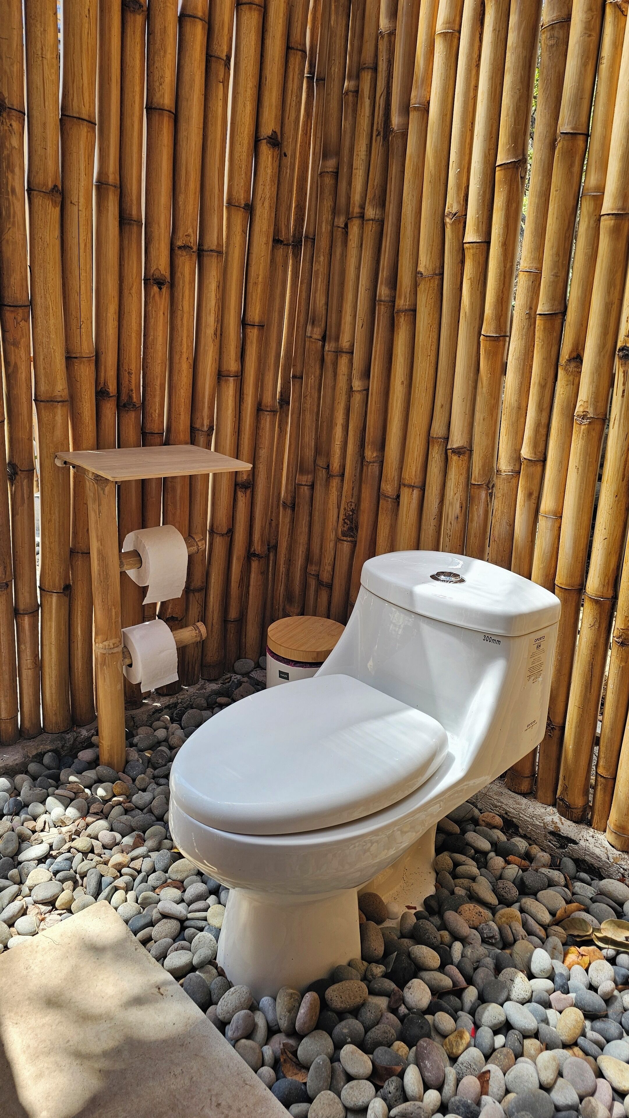 A toilet with a white seat and lid, positioned on a bed of pebbles, against a bamboo wall.