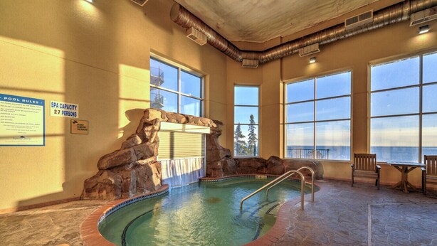 An indoor pool with a stone wall and a waterfall feature, surrounded by large windows and a stone floor. - Two Harbors Superior Lakefront 2BR | Pool | Hot Tub & Sauna | EV Charger (Two Harbors)