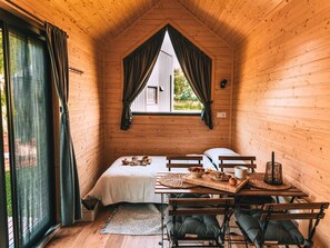 Una cabaña acogedora con cama en el altillo, una mesa de comedor con sillas y un ventanal con cortinas.