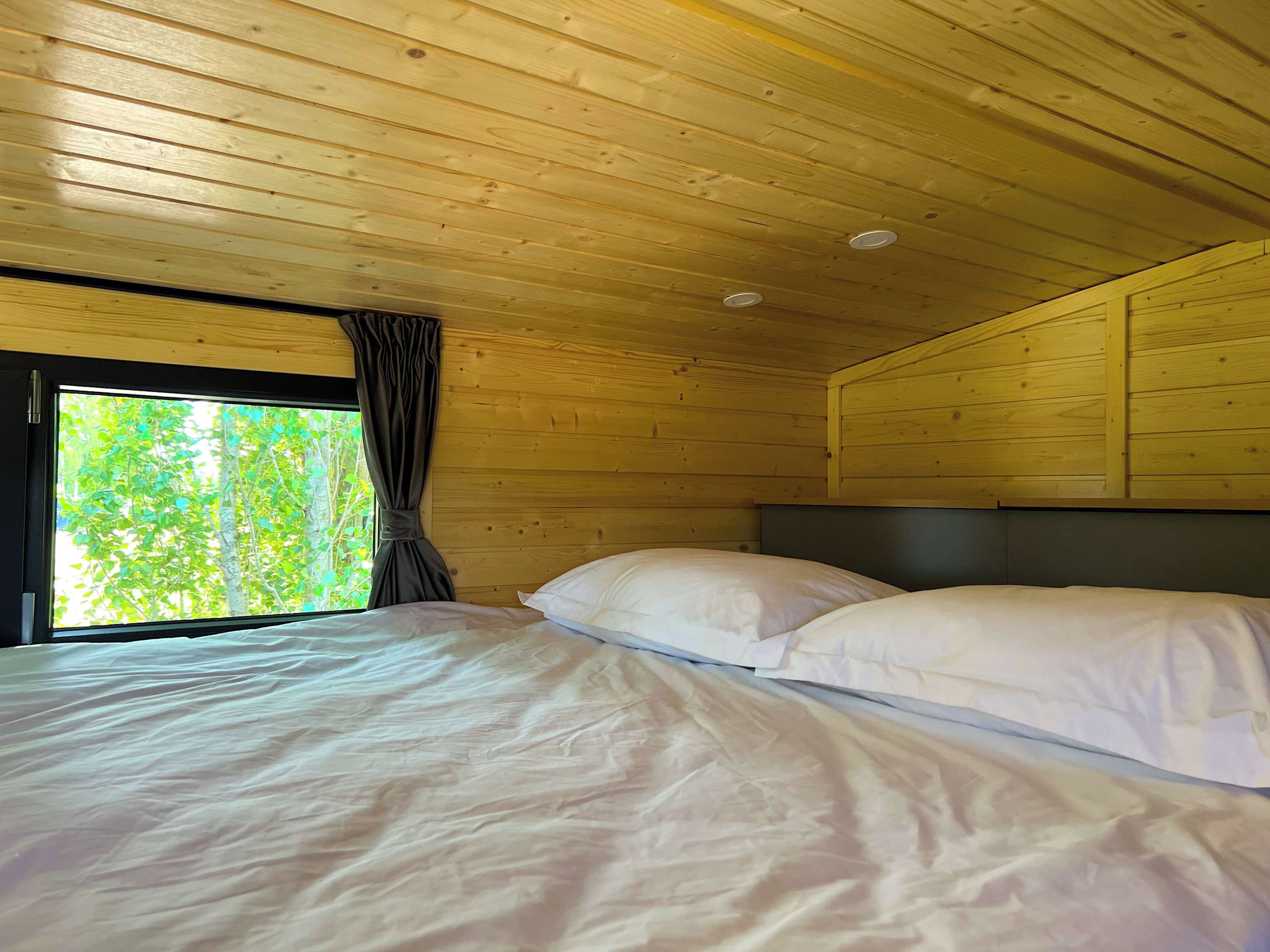 A wooden cabin room with a bed, a window with curtains, and a view of greenery outside.