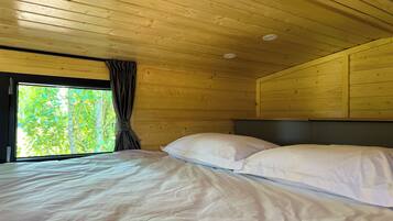 A wooden cabin room with a bed, a window with curtains, and a view of greenery outside.